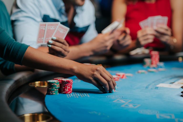Close-up of a hand placing betting chips on a casino table with other players holding playing cards in the background, implying a tense poker game.